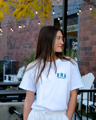 Woman wearing a white t-shirt with 'BNA' logo outdoors.
