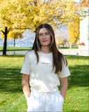 Person wearing a white t-shirt with text in an outdoor setting with trees and grass.