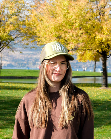 Person wearing a cap with 'BNA' on it in a park with trees and water in the background